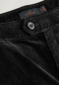 Black corduroy trousers with a textured surface, featuring a round grey button and a navy tag marked "NEXT SIGNATURE."