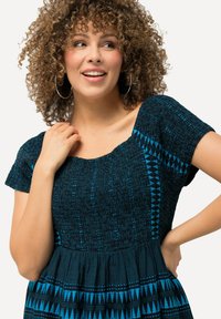 Musta ja sininen kuviollinen mekko, jossa on teksturoitu vartalo-osa, lyhyet hihat ja avara hame, jossa on geometrisia kuvioita.