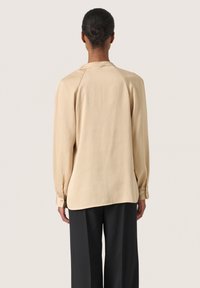 Beige langærmet bluse lavet af tekstureret stof, med rund halsudskæring og knaplukninger ved ærmerne, parret med løse sorte bukser.