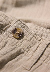 Beige textiel met een visgraatpatroon, met daarboven een ronde knoop met twee gaten boven een gestikte naad.