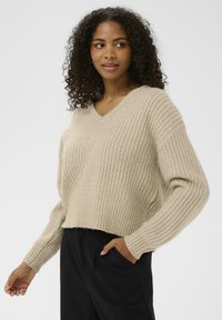 Beige knitted sweater with a V-neck, cropped design, and ribbed texture. Long sleeves and a relaxed fit complement its casual style.