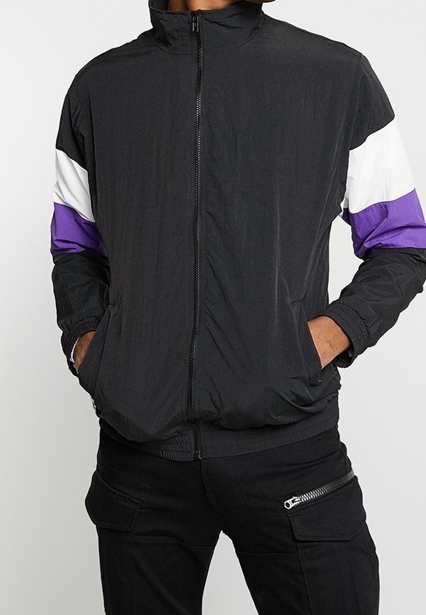 Veste noire zippée avec des bandes blanches et violettes sur les manches, portée avec un pantalon noir doté d'une poche zippée, mains dans les poches de la veste.