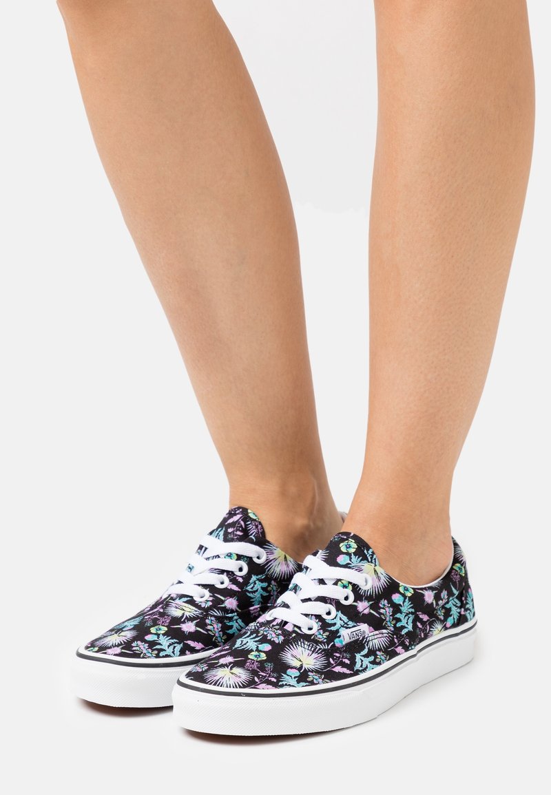 Baskets en toile noire avec un motif floral en violet et vert, équipées de lacets blancs et d'une semelle blanche.