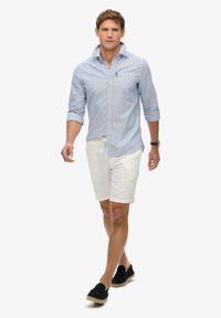 Blau-weißer gestreifter Button-up-Hemd, weiße Shorts und schwarze Slipper. Das Modell geht selbstbewusst und präsentiert lässige Sommerbekleidung.