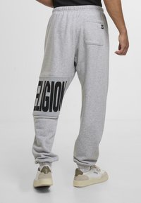 Sivé teplákové nohavice s veľkým čiernym potlačou "RELIGION" na ľavom stehne, elastický pás a zadná vrecko. Hladká textúra a voľný strih.
