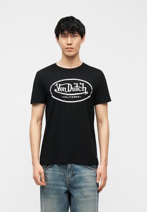 Mlad moški s črnimi lasmi, oblečen v črno majico z logotipom Von Dutch California in modre kavbojke, stoji pred enobarvnim sivim ozadjem.