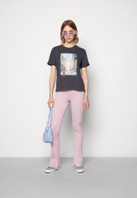 Camiseta cinza escura com um gráfico de paisagem urbana, emparelhada com calças flare rosa claro com detalhe texturizado e uma bolsa azul clara. O modelo usa óculos de sol roxos.