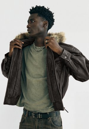 Junger Mann richtet eine braune Lederjacke mit Fellkragen, trägt ein grünes T-Shirt, eine Kette, Ringe und einen schwarzen Gürtel mit silbernen Ösen.