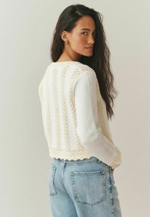 Moteris su ilgais tamsiais plaukais, dėvinti auksinius lankinius auskarus, kreminės spalvos tekstūruotą megztinį ir šviesiai mėlynas džinsines kelnes, stovinti priešais paprastą foną.