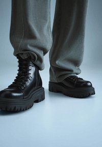 Černé kožené bojové boty s předním šněrováním, masivní gumovou podrážkou a zubatými okraji, doplněné šedými zúženými kalhotami.