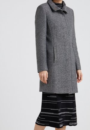 Femme portant un manteau gris texturé mi-long sur une jupe noire à rayures horizontales blanches, debout devant un fond uni.