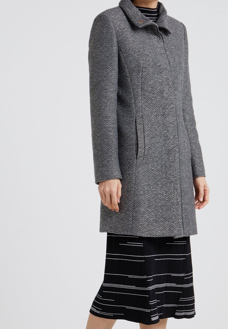 Femme portant un manteau gris texturé mi-long sur une jupe noire à rayures horizontales blanches, debout devant un fond uni.