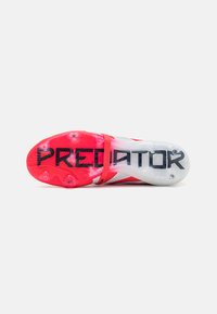 Červené a biele futbalové kopačky s textúrovaným zvrškom, spevneným páskom a priehľadnou podrážkou, na ktorej je zobrazené logo "PREDATOR" a tvarované kolíky.