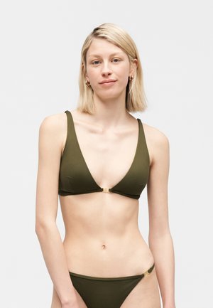 Lühikeste blondide juustega naine, kes kannab oliivrohelise värvi bikiine kuldsete kinnitustega, seisab heledal, ühtlasel taustal ja vaatab otse ette.