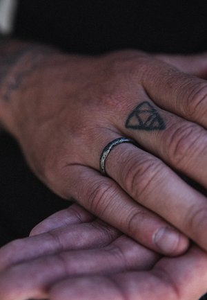Silver ring with a textured surface worn on a hand, displaying a tattoo of a triangle shape on the finger. Skin shows natural texture.