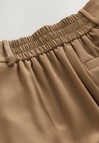 Tissu beige avec une ceinture élastique et des passants de ceinture sur un pantalon, montrant des coutures détaillées et une texture lisse.