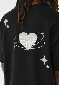 Zwarte T-shirt met een lichtgrijze hartgrafiek en de tekst "PROJECTX PARIS", omringd door cirkelvormige accenten en kleine sterdetails.