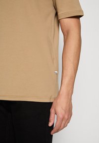Chemise à manches courtes beige en tissu doux, dotée d'une petite étiquette d'accent au bas et d'un col rond, portée avec un pantalon noir.