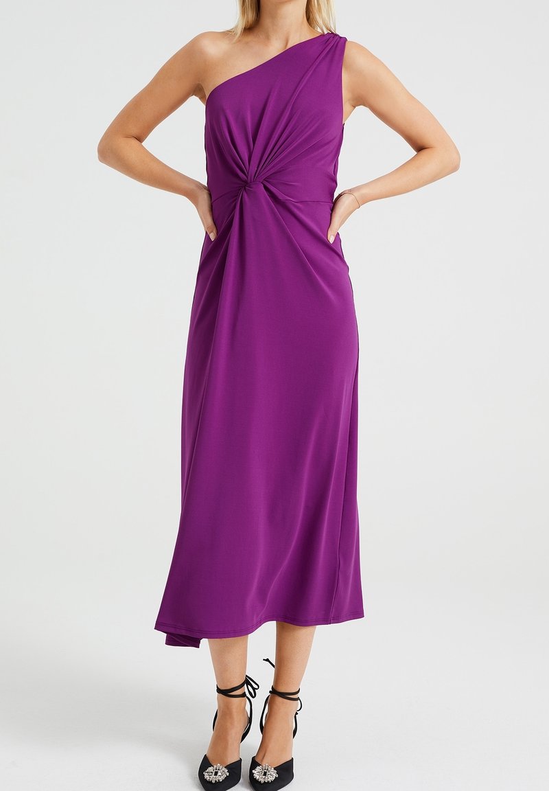 Fioletowa sukienka na jedno ramię z detalem w postaci węzła w pasie, wykonana z gładkiego materiału. Modelka ma na sobie czarne buty z ozdobnymi akcentami.