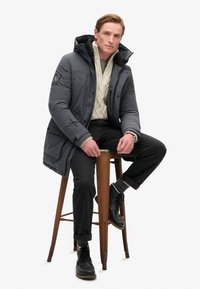 Doudoune grise avec capuche, arborant un patch logo, portée par-dessus un pull en maille torsadée crème. Pantalons noirs et bottes à lacets avec des chaussettes colorées.