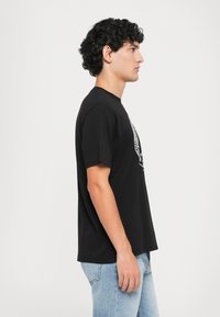 Joven con cabello rizado, vistiendo una camiseta gráfica negra y jeans claros, de pie de perfil contra un fondo blanco simple.
