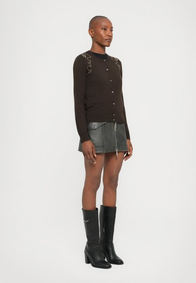 Bruine cardigan met knoopsluiting, met decoratieve borduursels op de schouders, gecombineerd met een grijze leren rok en zwarte kniehoge laarzen.