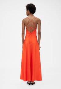 Maxi šaty z jasnej oranžovej saténovej látky s hlbokým výstrihom na chrbte, ktorý zdobia prekrížené remienky a zaväzovanie na mašľu. Hladká textúra a prúdiaca silueta.