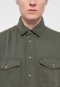 Olive green button-up shirt made of cotton, featuring two breast pockets, a collar, and brown button accents. Textured fabric.
