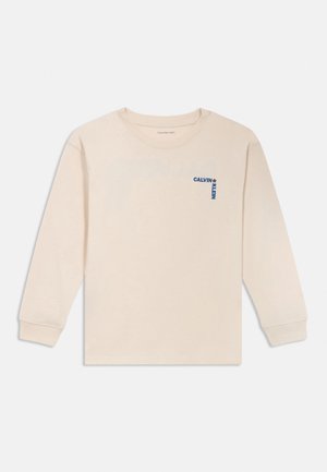 Kremowa koszula Calvin Klein z długim rękawem, z niebieskim pionowym napisem „CALVIN KLEIN” i gwiazdką na lewej piersi, z okrągłym dekoltem.