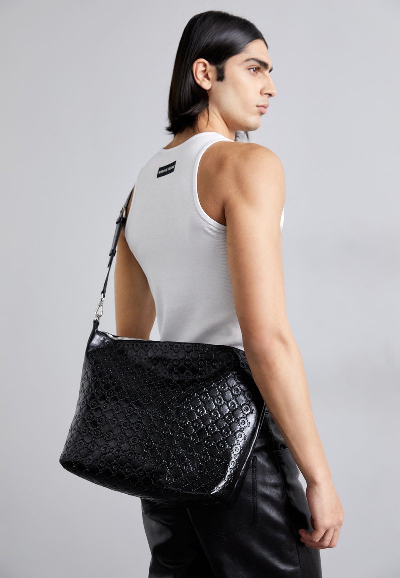 Black leather shoulder bag with a textured pattern, featuring a glossy finish and adjustable strap, being held by a person wearing a white tank.