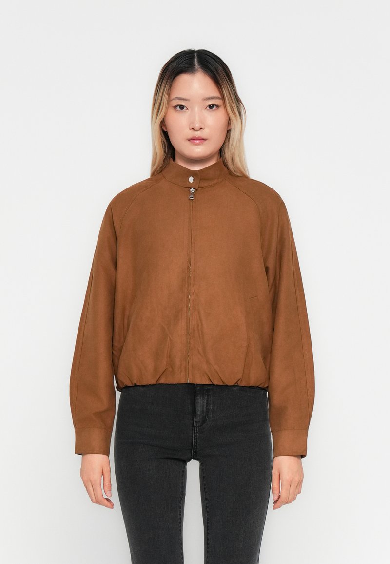 Kvinde med glat ombre hår iført en brun lynlåsjakke med krave og sorte højtaljede jeans, stående mod en ensfarvet hvid baggrund.