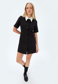 Vestido negro de manga corta con cuello blanco, cierre de botones en la parte delantera, dos bolsillos y textura suave. Corte ajustado, por encima de la rodilla.