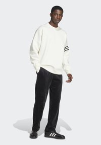 Sweatshirt blanc aux épaules tombantes avec des rayures noires sur les manches, associé à un pantalon en velours noir et des baskets noires brillantes.