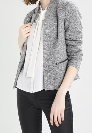 Femme portant un blazer gris texturé sur un chemisier blanc à col noué et un pantalon noir ajusté, debout devant un fond uni.