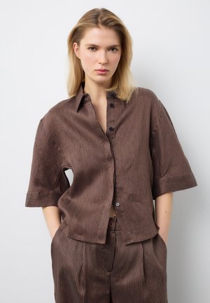 Frau trägt locker sitzendes braunes Nadelstreifenhemd mit Ellenbogenlangen Ärmeln und passende Hose, steht mit den Händen in den Taschen vor weißem Hintergrund.