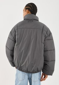 Graue Pufferjacke mit hohem Kragen, elastischem Saum und Steppdetails. Verfügt über ein Logo auf der Rückseite, kombiniert mit hellblauen Jeans.