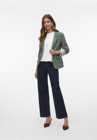Lichtgroene blazer met opgerolde mouwen, witte top en donkerblauwe wijde jeans; bruine hakken met een spitse neus en decoratieve details.