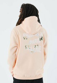 Sudadera melocotón con un diseño gráfico floral blanco y el texto "PROJECT X PARIS GALERIE" en la parte posterior. Tela suave, corte relajado.