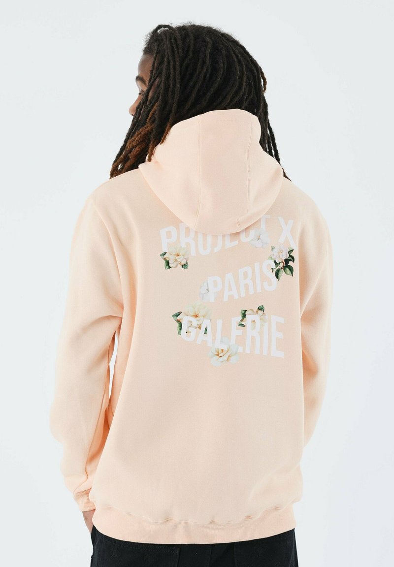 Sudadera melocotón con un diseño gráfico floral blanco y el texto "PROJECT X PARIS GALERIE" en la parte posterior. Tela suave, corte relajado.