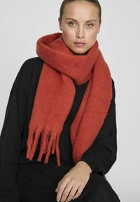 Écharpe en laine rouge avec des bords frangés, dotée d'une finition douce et texturée. Portée librement autour du cou sur un haut noir.
