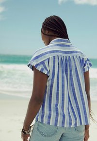Kurzärmliges, lässiges Hemd mit blauen und weißen vertikalen Streifen, mit einem Rückenfalte und Kragen. Getragen mit Denim-Shorts.