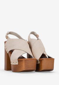 Sandalias de plataforma de ante beige con diseño de tiras cruzadas, tacones de bloque de madera y tachuelas de metal. Incluye una correa ajustable en el tobillo para un mejor ajuste.