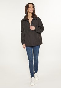 Schwarze Zip-Jacke mit hohem Kragen, elastischen Bündchen und lockerer Passform, kombiniert mit blauen Jeans und weißen Turnschuhen. Schlichte, ungeschmückte Gestaltung.