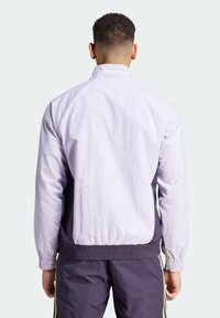 Chaqueta cortavientos de color púrpura claro con puños elásticos y un acento púrpura oscuro en los lados. Textura suave, diseño minimalista, vista trasera.