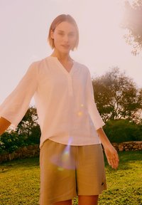 Femeie stând în aer liber pe iarbă, purtând o bluză albă cu mâneci lungi și pantaloni scurți bej, cu copaci luminați de soare și un zid de piatră în fundal.