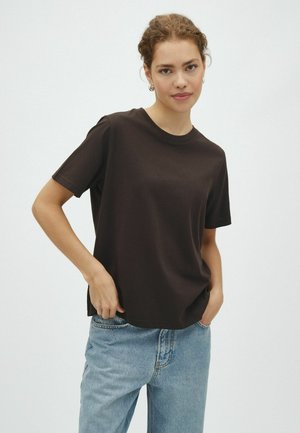 Donna con capelli ricci che indossa una t-shirt nera a maniche corte e jeans azzurri, in piedi con le mani in tasca davanti a uno sfondo uniforme.