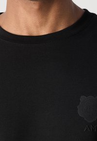 Detailní záběr na osobu s černým tričkem s kulatým výstřihem, na němž je malá, texturovaná černá tygří emblema a částečně viditelné písmena na hrudi.