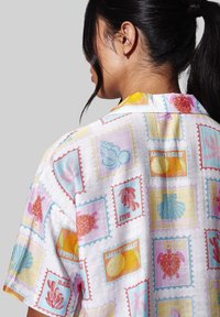 Mujer con cabello negro recogido en una coleta, que lleva una camisa colorida con estampados de tortugas marinas, conchas, coral y el texto "Costa de Amalfi".