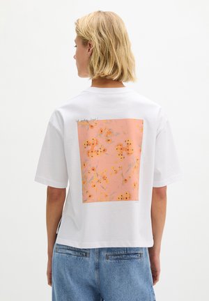 Blonde Person trägt ein weißes T-Shirt mit einem großen pfirsichfarbenen Blumenmuster auf dem Rücken, kombiniert mit blauen Jeans aus Denim.