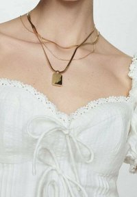 Colliers superposés en or avec un pendentif rectangulaire, présentés sur un cou vêtu d'un haut blanc texturé avec des accents en dentelle.